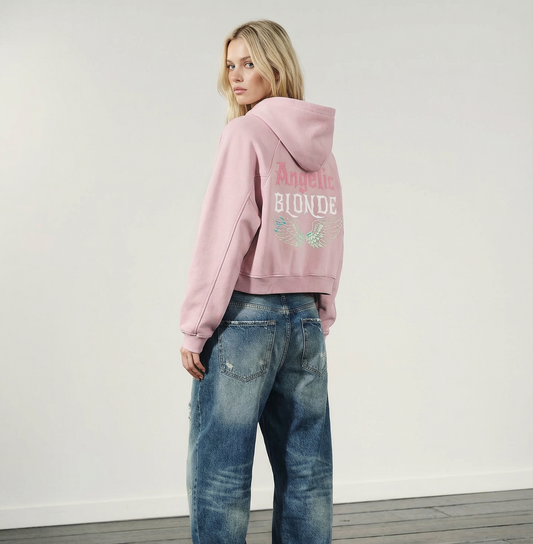 Back view of a model wearing of a blush-pink cropped hoodie with hood up, featuring “Angelic Blonde” New Rocker font text and iridescent angel wings graphic printed across the back on a white background.