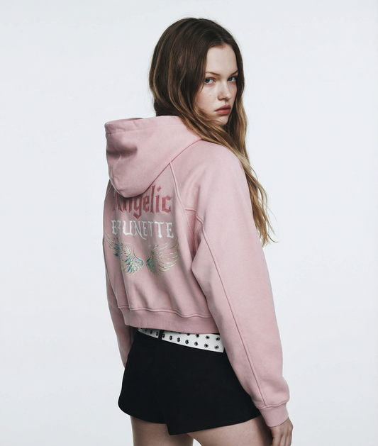 model wears a a pale pink cropped hoodie with hood up, featuring “Angelic Brunette” New Rocker font text and iridescent angel wings graphic printed across the back on a white background.