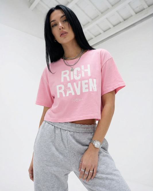 raven haired model wears a pink crop top with a relaxed fit, short sleeves, and a wide crew neckline. The front features bold white distressed lettering reading “RICH RAVEN” with the RedBlonde signature logo underneath in black