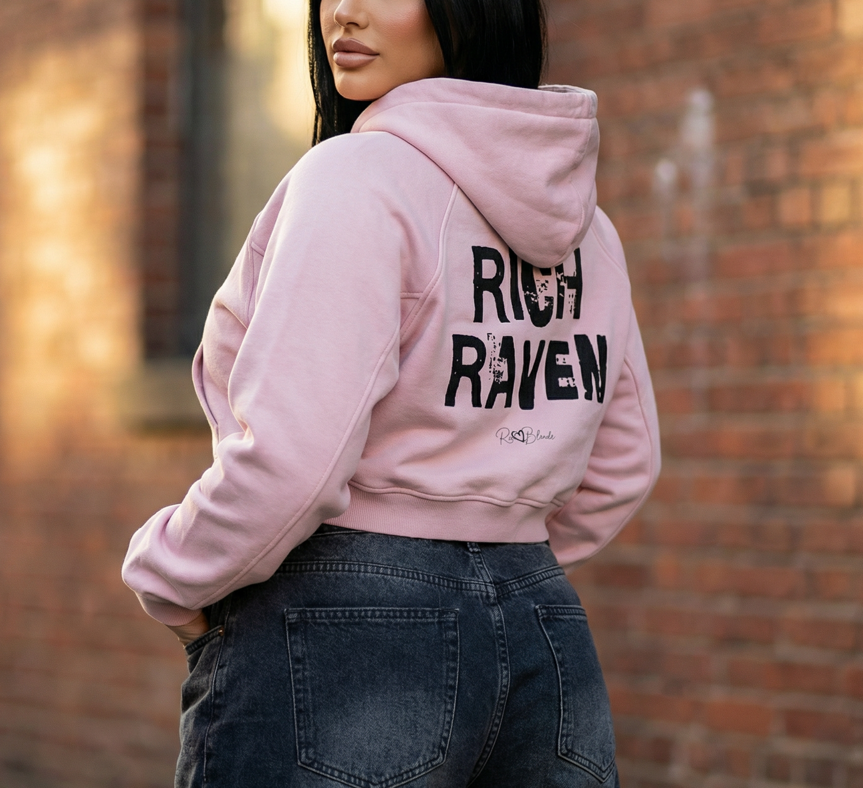 model wears  a cropped snow-wash pale pink hoodie with bold black “RICH RAVEN” Ed Gein font and the RedBlonde signature logo printed below.