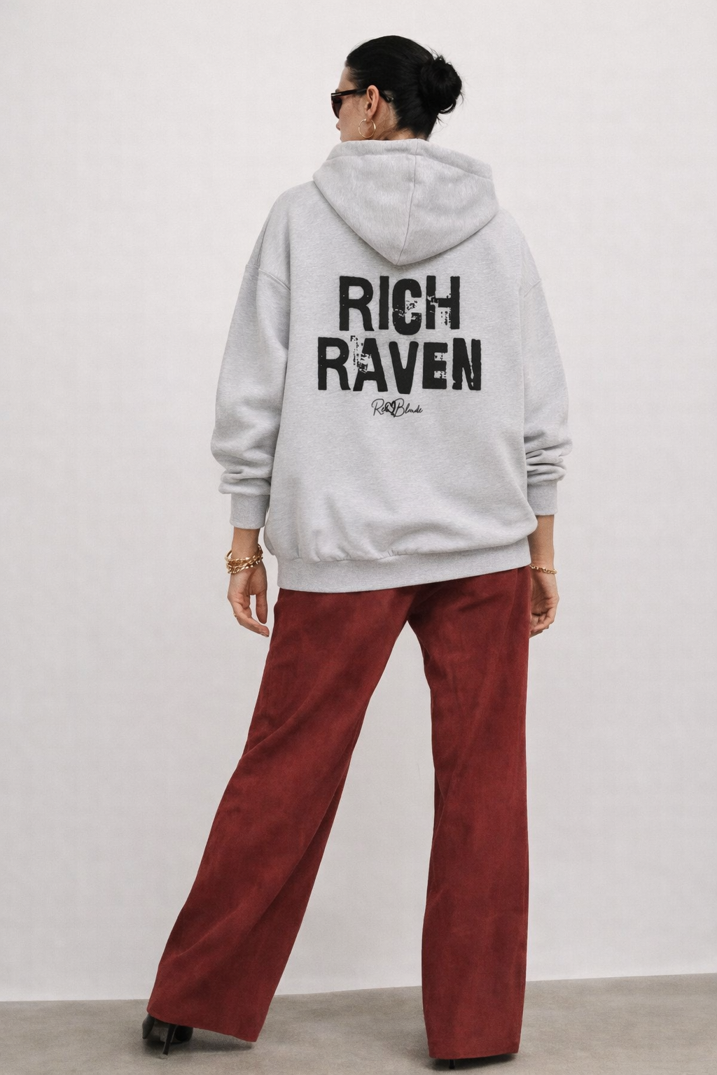 model wears grey hem to hood zip-up hoodie with cropped oversized fit and ribbed cuffs/hem, featuring large distressed black “RICH RAVEN” Ed Gein text across the back with a small “RedBlonde” script logo beneath.