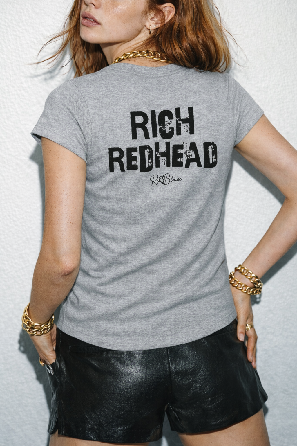 model wears a  grey slightly fitted women’s henley T-shirt with short sleeves and a gently curved hem. Centered on the upper back is bold distressed black Ed Gein text reading “RICH REDHEAD” with a small RedBlonde signature script logo beneath it. 