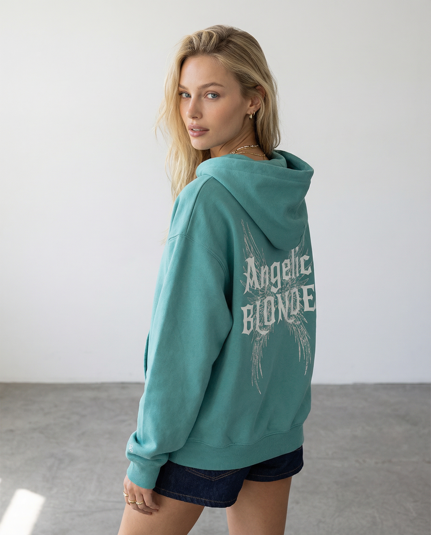 model wears Green Teal oversized fleeced hoodie with a back print featuring white gothic “Angelic Blonde” text layered over large angel wing artwork.