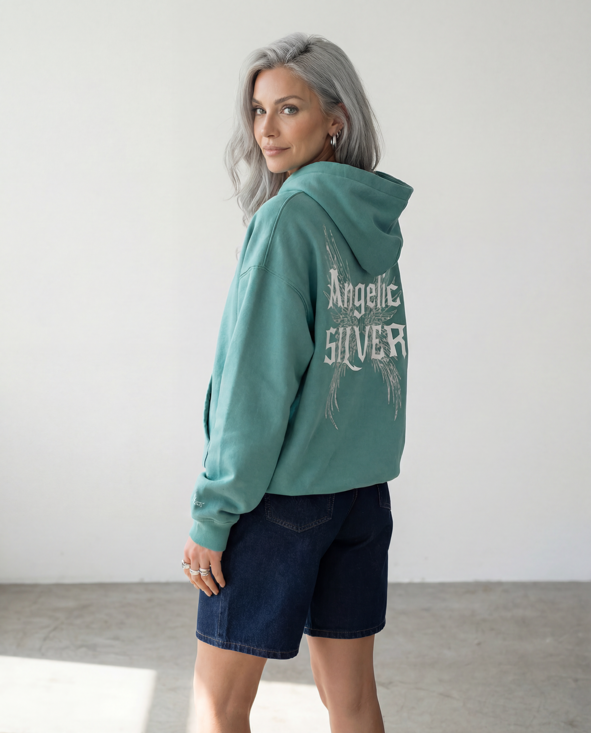 model wears Teal oversized fleeced hoodie with a back print featuring white gothic “Angelic Silver” text layered over large angel wing artwork.