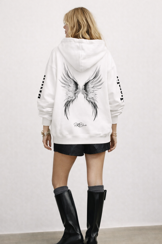 model wears a white hoodie featuring large silver angel wings printed across the back, with black RedBlonde signature logo underneath. Each sleeve features gothic New Rockstar font graphic text  one sleeve says 'Angelic' and the other 'Blonde' 