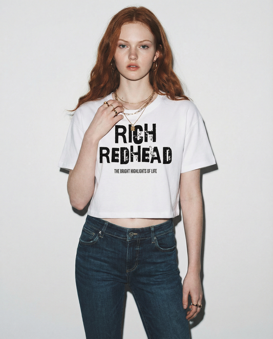 Redhead wears A white women’s crop top with a relaxed fit, short sleeves, and a wide crew neckline. The front features bold black distressed lettering reading “RICH REDHEAD” with the tagline “THE BRIGHT HIGHLIGHTS OF LIFE” in smaller text below. 