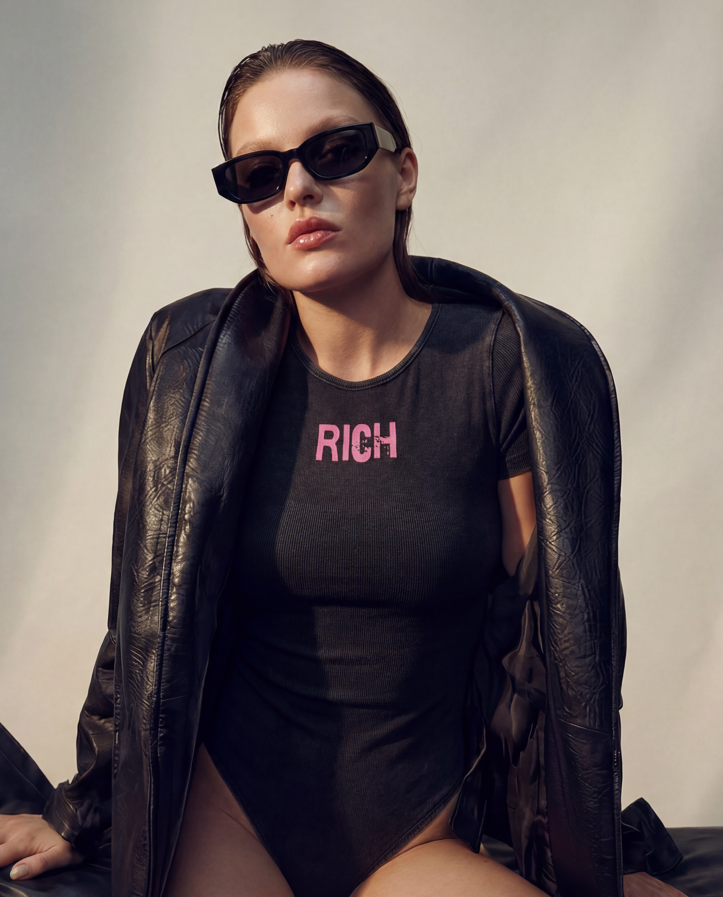 model wears a Washed charcoal ribbed short sleeve bodysuit with a crew neckline and pink 'RICH'  in Ed Gein font graphic at the chest.