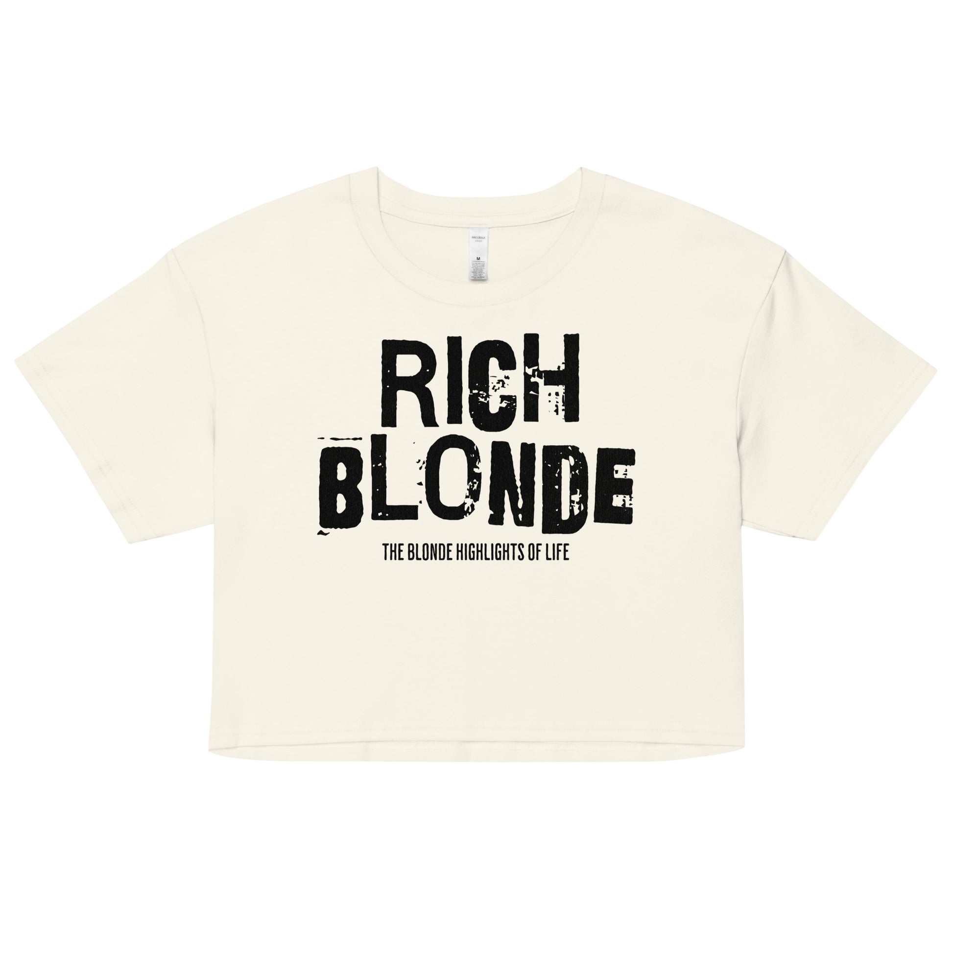 A natural women’s crop top with a relaxed fit, short sleeves, and a wide crew neckline. The front features bold black distressed lettering reading “RICH BLONDE” with the tagline “THE BLONDE HIGHLIGHTS OF LIFE” in smaller text below. The minimalist design contrasts sharply against the soft cream fabric