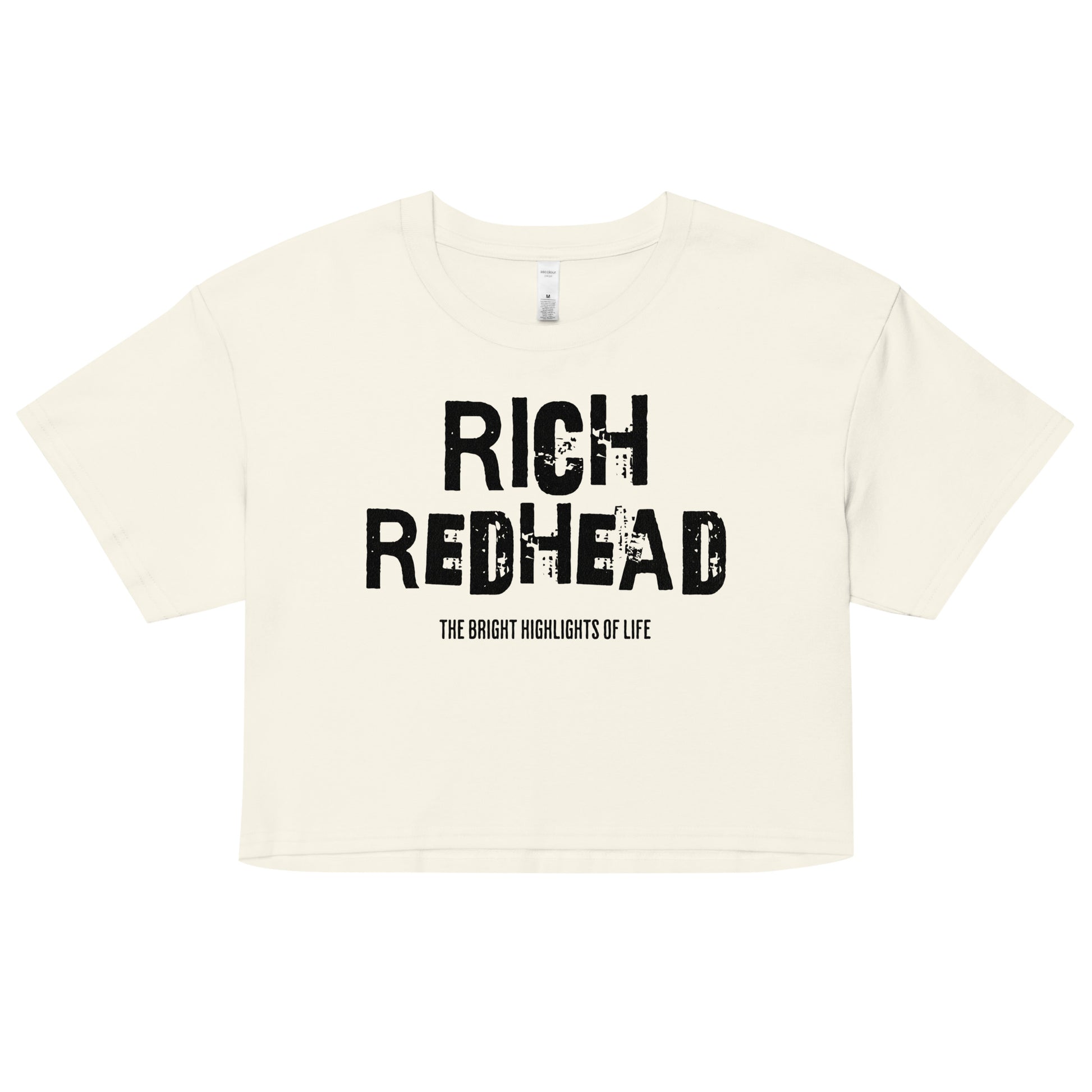 A natural women’s crop top with a relaxed fit, short sleeves, and a wide crew neckline. The front features bold black distressed lettering reading “RICH REDHEAD” with the tagline “THE BRIGHT HIGHLIGHTS OF LIFE” in smaller text below. The minimalist design contrasts sharply against the soft cream fabric
