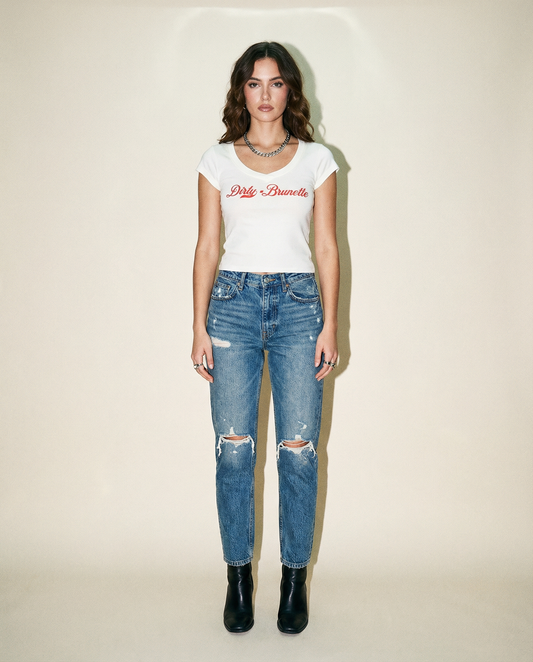 model wears A fitted white V-neck T-shirt. It has short sleeves and a red Milkstore 01 script graphic across the chest reading “Dirty-Brunette.” 