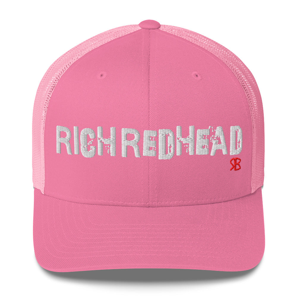 pink trucker cap with the bold distressed text “RICH REDHEAD” embroidered in white across the front, and a small red “RB” logo detail beneath the text. The cap has a structured front panel and curved brim