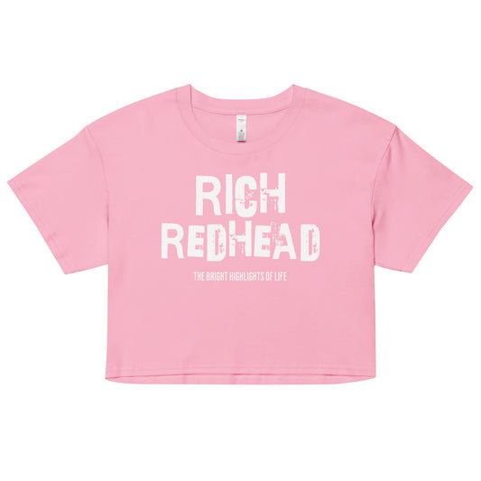 A pink women’s crop top with a relaxed fit, short sleeves, and a wide crew neckline. The front features bold black distressed lettering reading “RICH REDHEAD” with the tagline “THE BRIGHT HIGHLIGHTS OF LIFE” in smaller text below. 