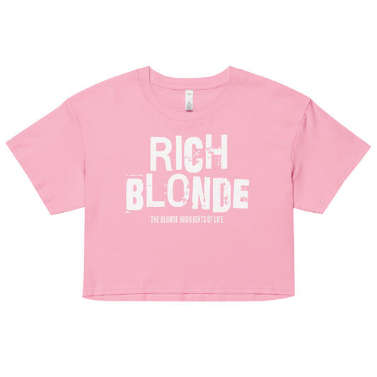 A pink women’s crop top with a relaxed fit, short sleeves, and a wide crew neckline. The front features bold white distressed lettering reading “RICH BLONDE” with the tagline “THE BLONDE HIGHLIGHTS OF LIFE” in smaller text below. 