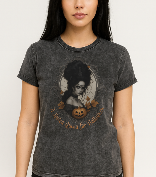 woman with black hairs wears a Black acid-wash unisex T-shirt featuring a gothic-style illustration of a dark-haired woman with an elaborate beehive hairstyle, framed by ornate scrollwork, autumn leaves, and a carved pumpkin. The design is captioned with the text ‘A Raven Queen for Halloween’ in vintage Dark Angle lettering. Branded RedBlonde in red on inside neck and right sleeve