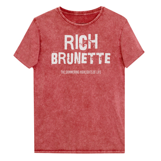 A red acid-wash unisex denim T-shirt with a distressed vintage look. The shirt features the bold white text “RICH BRUNETTE” in a weathered Ed Gein font, with the tagline “THE SHIMMING HIGHLIGHTS OF LIFE” in smaller uppercase letters below. The shirt has short sleeves, a crew neckline, and visible seam detailing for a rugged, casual style. Branded Redblonde on inside neck and right sleeve