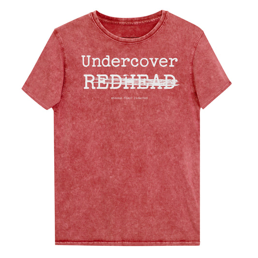 A red unisex denim-style t-shirt with a vintage washed finish. The front features bold white text reading “Undercover REDHEAD” partially struck through by a horizontal white bar. Beneath it, smaller text says “mission file // redacted” along with a subtle RedBlonde logo. The shirt has a classic crew neck and short sleeves with visible stitching for a rugged look. Branded RedBlonde London in red on inside neck and right sleeve