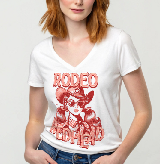 redhead wears a Women's white V-neck t-shirt featuring a bold red vintage illustration of a cowgirl with heart-shaped sunglasses and pigtails, wearing a wide-brimmed cowboy hat and scarf. Text reads "RODEO REDHEAD" in large Western-style lettering. Branded RedBlonde on inside neck and right sleeve.