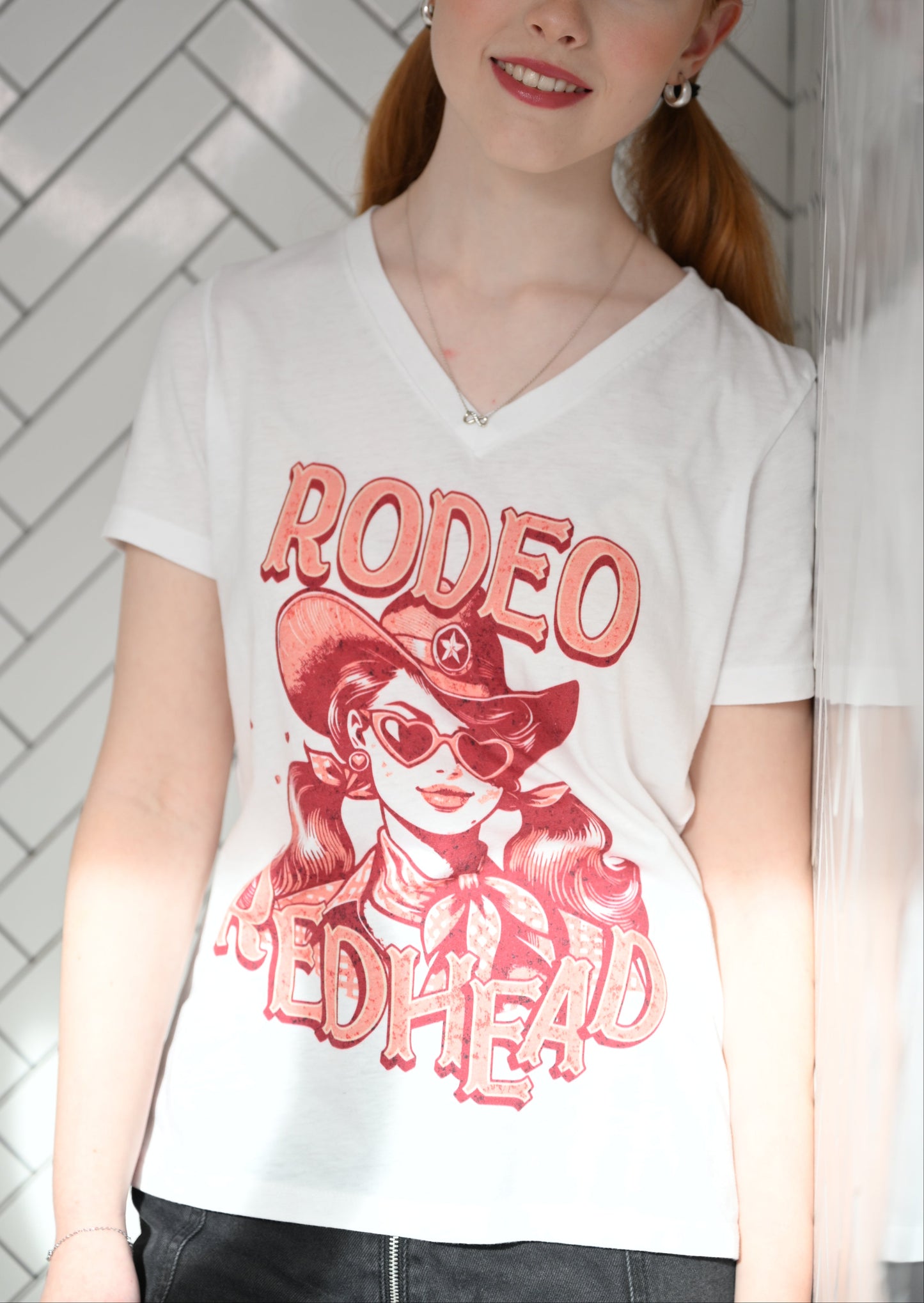 redhead wears a Women's white V-neck t-shirt featuring a bold red vintage illustration of a cowgirl with heart-shaped sunglasses and pigtails, wearing a wide-brimmed cowboy hat and scarf. Text reads "RODEO REDHEAD" in large Western-style lettering. Branded RedBlonde on inside neck and right sleeve.