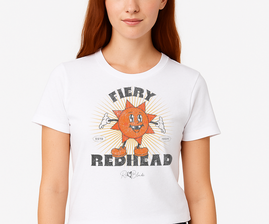 redhead wears a White unisex crew neck t-shirt with a bold retro graphic of a smiling orange cartoon sun character, surrounded by radiant sunbeams. The text reads ‘Fiery Redhead’ in distressed black Belzebuth lettering with small details including ‘ESTD 1994’ and the RedBlonde signature logo beneath."