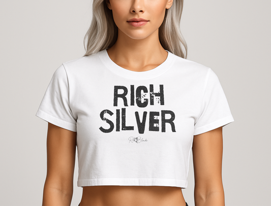 silver haired model wears  a white crop top with a relaxed fit, short sleeves, and a wide crew neckline. The front features bold black distressed lettering reading “RICH SILVER” with the RedBlonde signature logo underneath in black