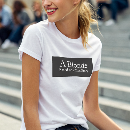 blonde model, sitting on stone steps, wears a white unisex relaxed fit t-shirt with the slogan 'A Blonde. Based on a True Story' in white text in a black box graphic 